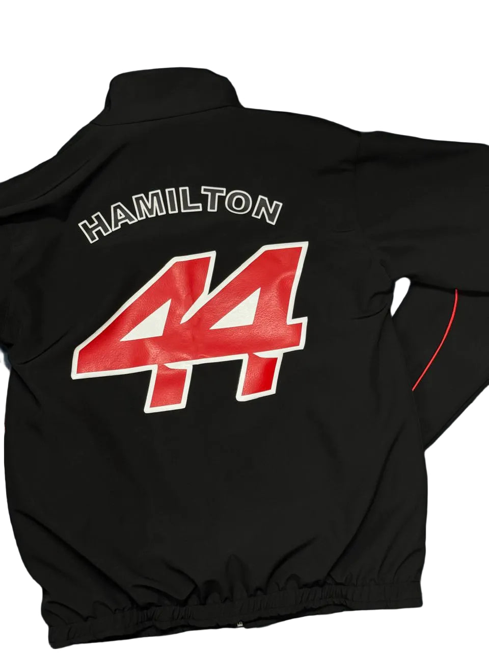 A black windbreaker jacket laid flat on a blue fabric surface, showing the back with 'HAMILTON' in white outlined letters above a large red number 44 with a white outline.