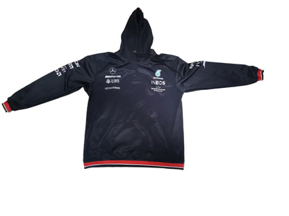 A black Mercedes Formula 1 team hoodie jacket with red and white stripes on the cuffs and hem, laid flat on a light gray tiled floor. The jacket features white sponsor logos and text, including Mercedes-Benz, F1, and Castore.