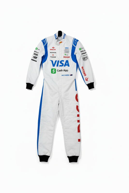 A white racing one-piece suit with blue and red sponsor logos, laid flat on a brown concrete floor. A bare foot is visible at the bottom left corner.