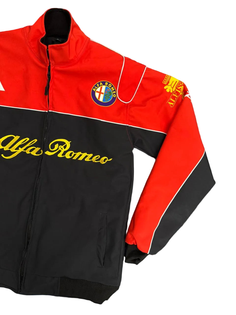 An Alfa Romeo track jacket laid flat on a blue fabric surface, featuring a red and black color-blocked design with yellow script lettering and team crests.