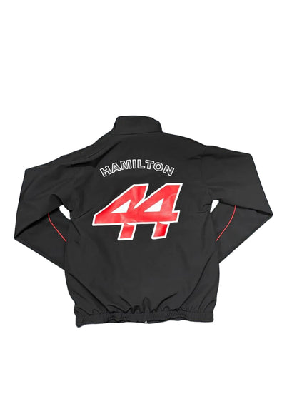 A black zip-up jacket laid flat on a blue sheet, showing the back with 'HAMILTON' and the number '44' in red and white. Bare feet are visible at the bottom edge, and a room floor and storage items are in the background.